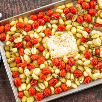 A rimmed sheet pan is full of roasted gnocchi, cherry tomatoes and a browned block of feta, with sprigs of thyme scattered throughout. A metal serving spoon is tucked into the tomatoes and gnocchi. A dish towel is nestled next to the pan.