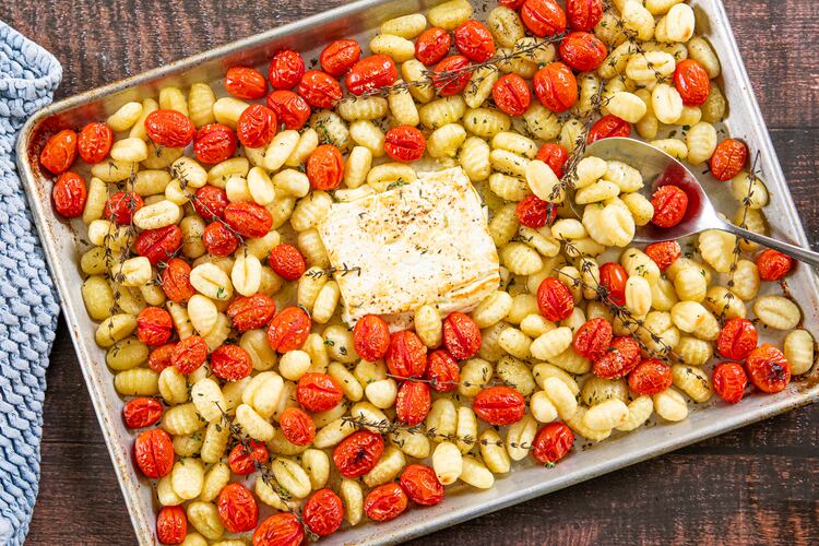 A rimmed sheet pan is full of roasted gnocchi, cherry tomatoes and a browned block of feta, with sprigs of thyme scattered throughout. A metal serving spoon is tucked into the tomatoes and gnocchi. A dish towel is nestled next to the pan.