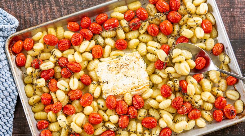 A rimmed sheet pan is full of roasted gnocchi, cherry tomatoes and a browned block of feta, with sprigs of thyme scattered throughout. A metal serving spoon is tucked into the tomatoes and gnocchi. A dish towel is nestled next to the pan.
