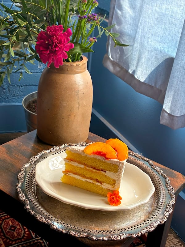 A slice of cake with white icing garnish with peaches stands next to a flower vase.