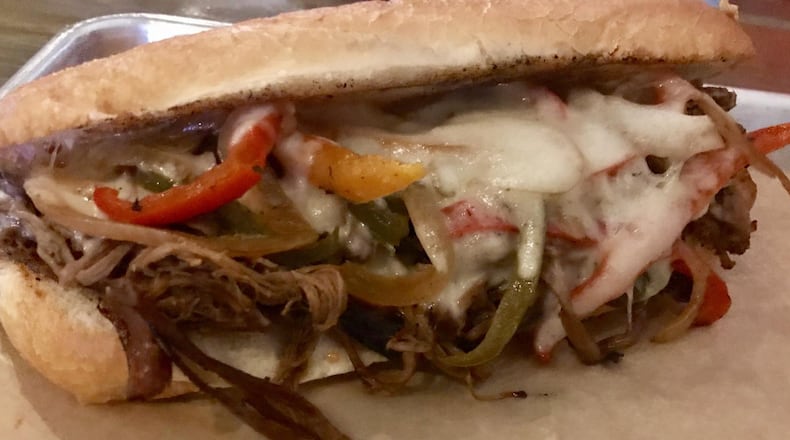 The Philly French Dip at Mouth of the South features smoked brisket, a colorful trio of grilled pepper slices and onions, gooey melted Swiss and provolone cheeses and Sriracha mayo on a toasted hoagie. The sandwich is served with au jus and comes with a side of chips and a pickle. LIGAYA FIGUERAS / LFIGUERAS@AJC.COM