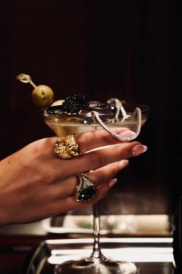A woman's jeweled hand holds a martini garnished with olives and spoonful of caviar.