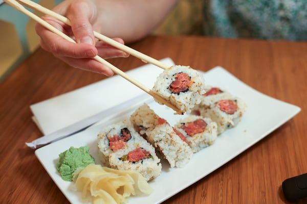 A hand picks up a piece of spicy tuna roll using chopsticks. The sushi is on a plate with wasabi paste and pickled ginger. The tuna roll has been cut into 8 pieces.