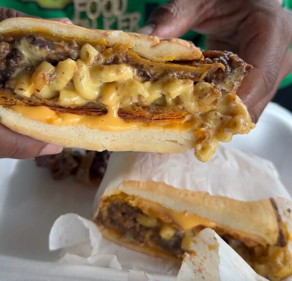 Hands present half of a sandwich to the camera. The sandwich is thick with a meat patty, braised oxtail and noticeably dripping, ooey gooey macaroni and cheese.