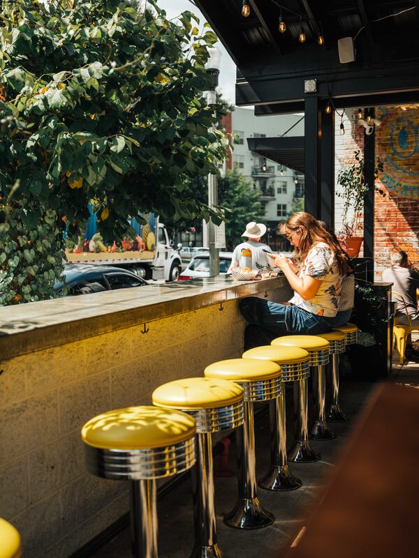 An outdoor patio with vintage barstools with bright sunlight flooding the space. One person sits on the far end of the bar/wall.