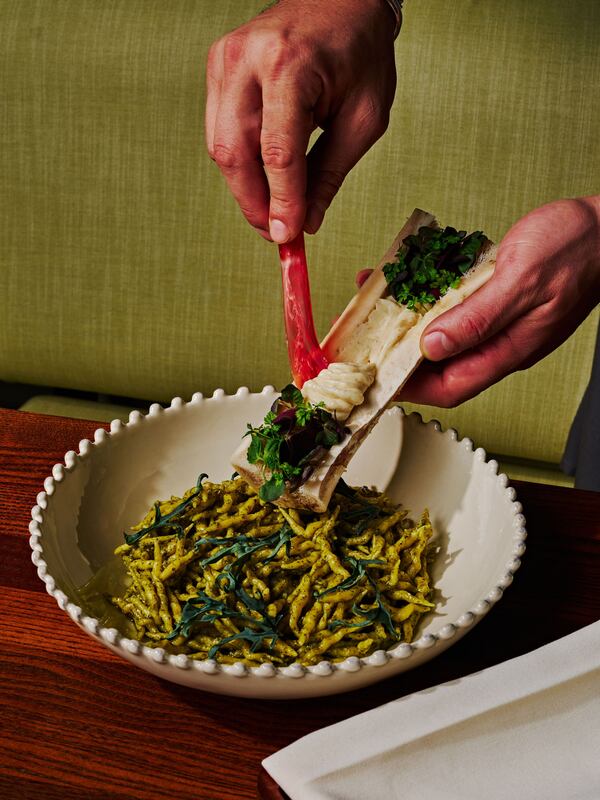A bowl of short, twisty pasta in a bright green sauce, with hands reaching in from the top of the frame using a spoon to scrape marrow butter out of a clean, white bone.