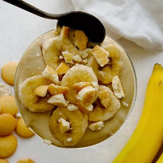 A glass of homemade banana pudding topped with sliced bananas and crushed vanilla wafers, with a spoon resting on top.
