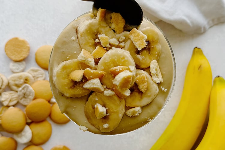 A glass of homemade banana pudding topped with sliced bananas and crushed vanilla wafers, with a spoon resting on top.