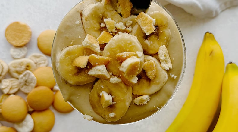 A glass of homemade banana pudding topped with sliced bananas and crushed vanilla wafers, with a spoon resting on top.