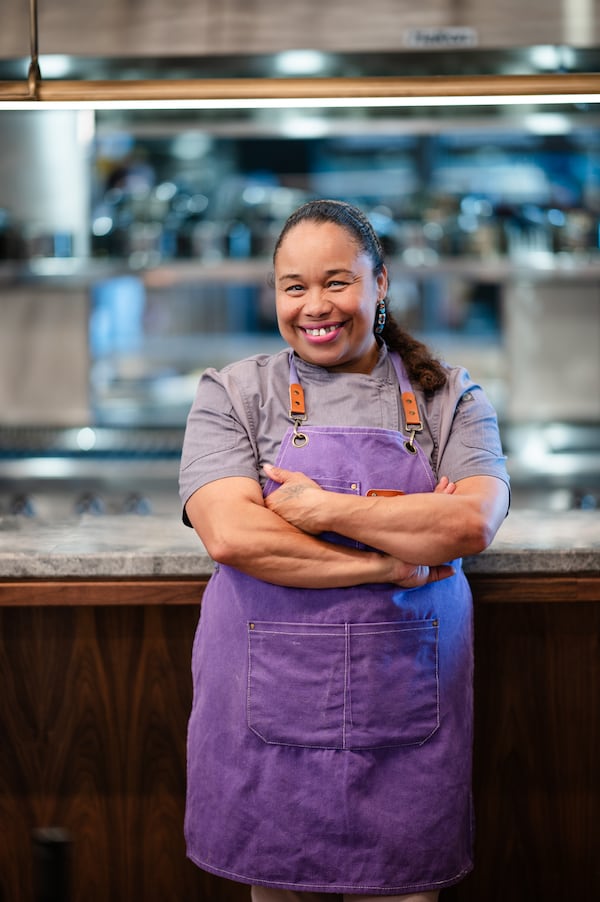 A woman chef in an apron stands in front of a kitchen counter facing the camera, smiling with her arms crossed.