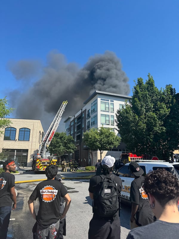 Several people in the foreground look away toward an apartment building which has a fire truck in front of it and smoke billowing from the top.