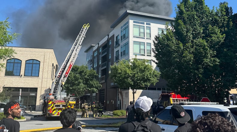 Several people in the foreground look away toward an apartment building which has a fire truck in front of it and smoke billowing from the top.