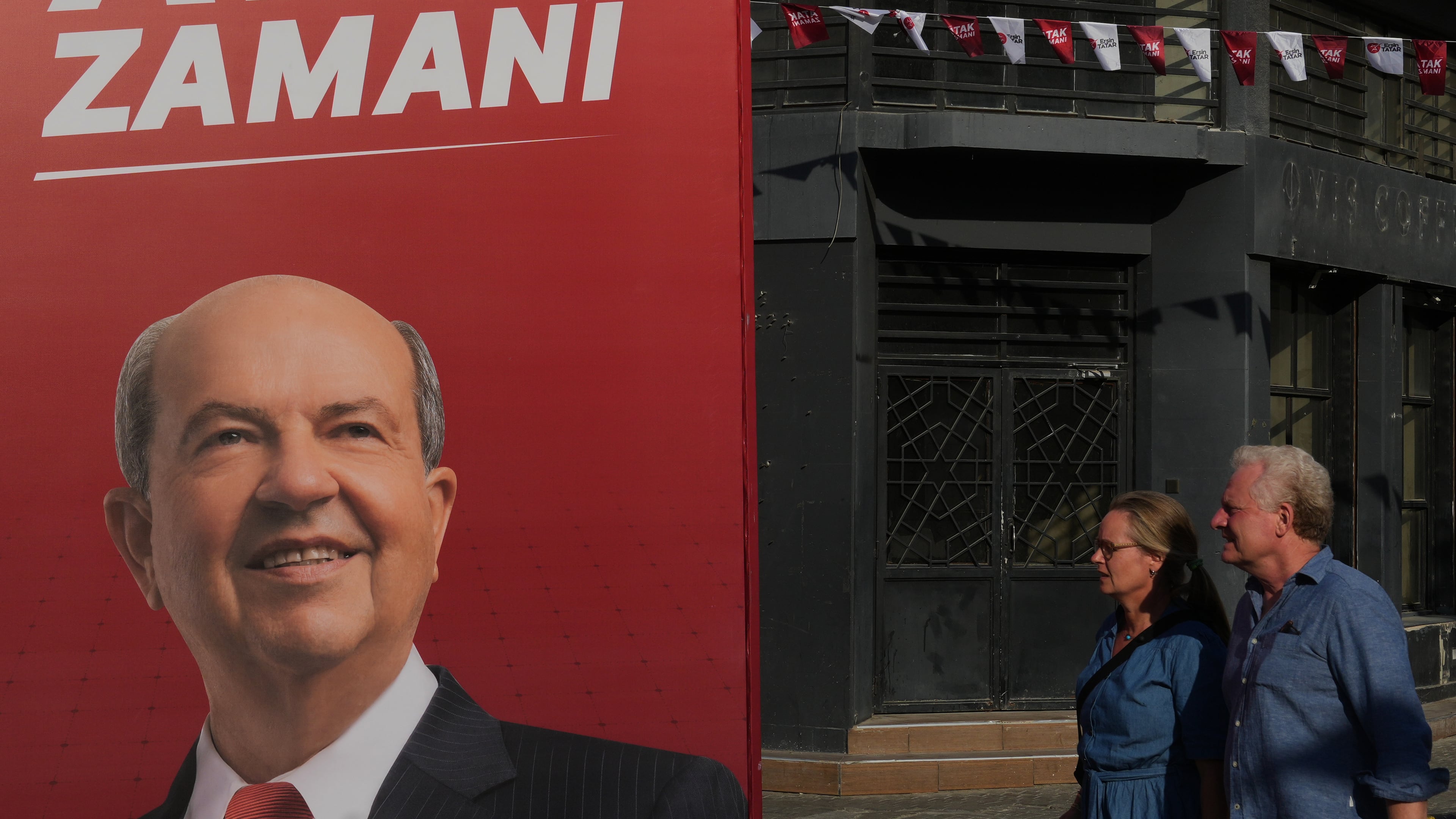 A couple walk past a campaign poster of Turkish Cypriot leader and candidate Ersin Tatar ahead of the Turkish Cypriot elections in the Turkish occupied northern part of divided Nicosia, Cyprus, Friday, Oct. 17, 2025. (AP Photo/Petros Karadjias)