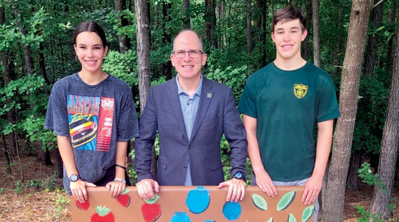 Faustina Barcena (left) stands with Adam Pomeranz, president and CEO of Annandale Village (center), and her brother, John, in front of a bench she created for the Suwanee assisted living facility. Courtesy