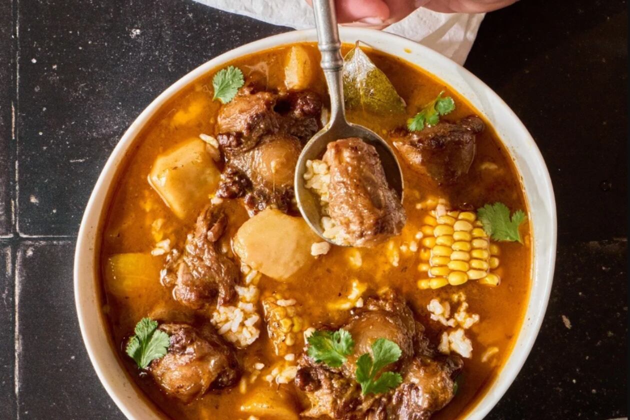 Overhead view of a bowl of sancocho with oxtails, corn, potatoes, root vegetables and rice in a rich tomato based broth. The stew is in a white bowl and is garnished with cilantro. A spoon lifts a piece of meat.