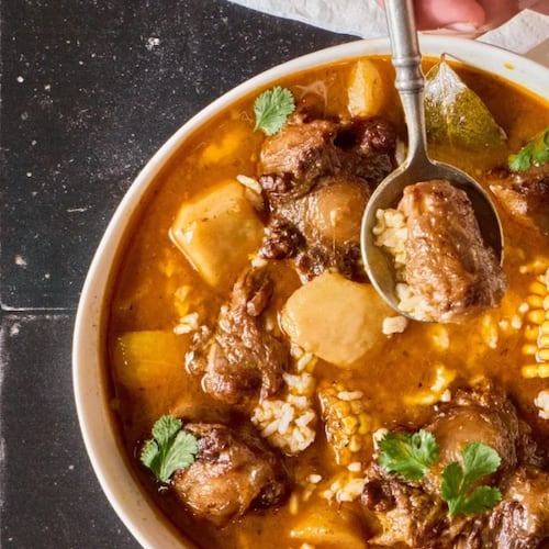 Overhead view of a bowl of sancocho with oxtails, corn, potatoes, root vegetables and rice in a rich tomato based broth. The stew is in a white bowl and is garnished with cilantro. A spoon lifts a piece of meat.
