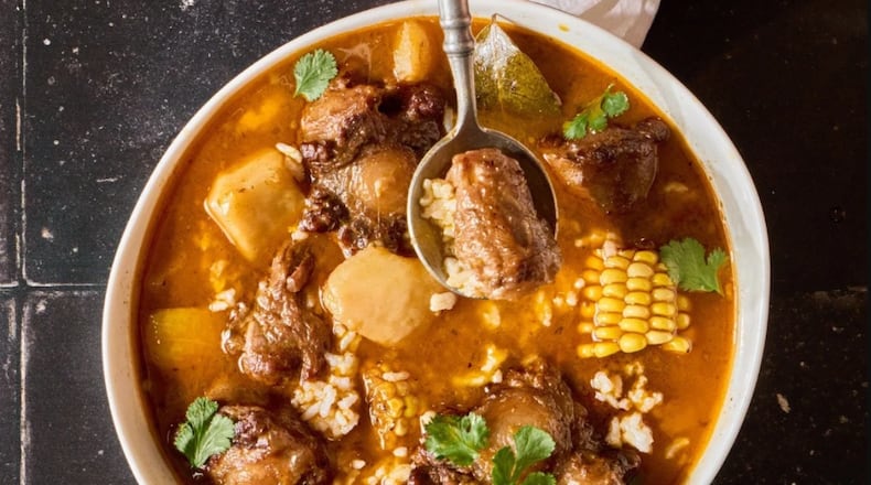 Overhead view of a bowl of sancocho with oxtails, corn, potatoes, root vegetables and rice in a rich tomato based broth. The stew is in a white bowl and is garnished with cilantro. A spoon lifts a piece of meat.