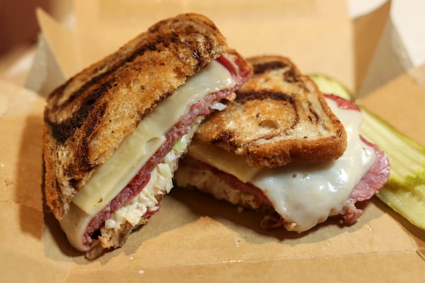 A pastrami Reuben on marbled rye bread is sliced in half to show the Swiss cheese and coleslaw inside. It is in a cardboard take-out container and accompanied by slices of deli pickles.
