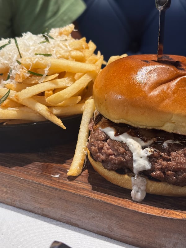 A burger and fries served on a wood plank. The thick burger has a drip of melted goat cheese and a smooth bun. A steak knife is stabbed into the center of the burger. The french fries are lightly golden with what appears to be a sprinkle of shredded Parmesan cheese on top.