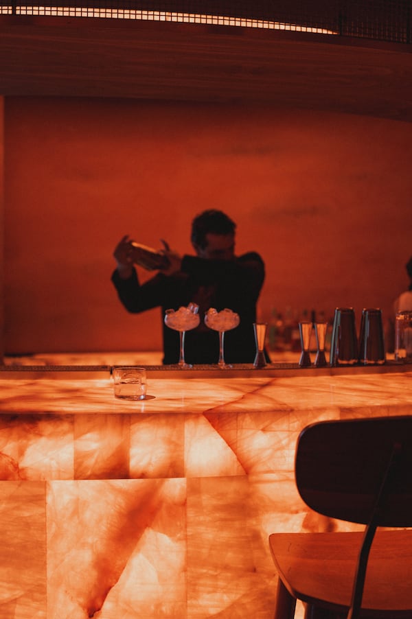 A bartender makes cocktails in a dim but warmly lit bar, with a stone bar lit from within in front of him and red stone walls behind him