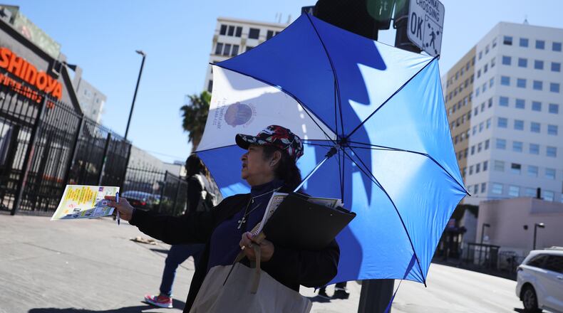 A woman covers herself from the sun while handing out fliers during an unseasonably hot day at MacArthur Park on Thursday, March 12, 2026, in Los Angeles. (AP Photo/Ryan Sun)