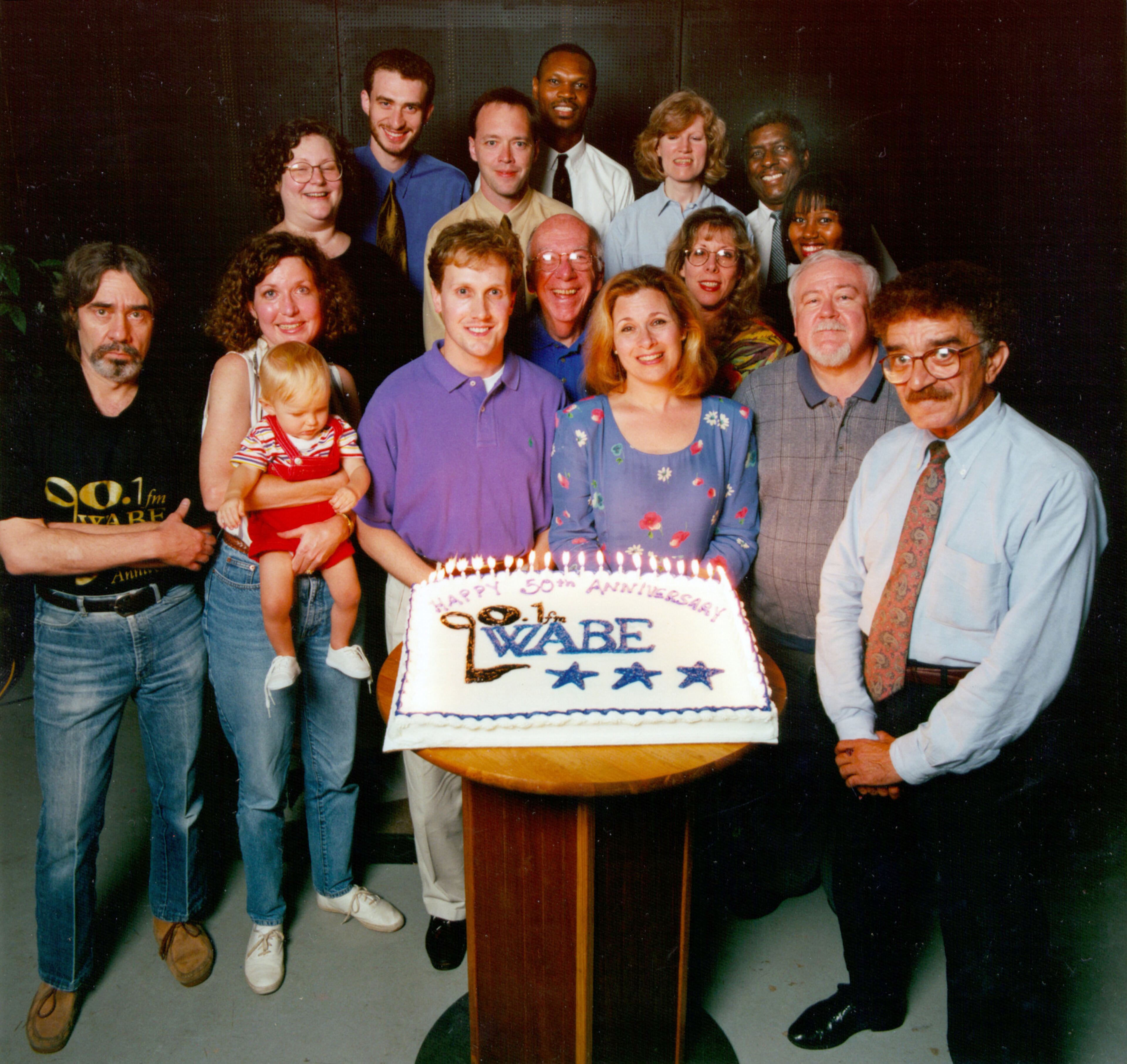Members of the WABE family celebrating an anniversary in 1998: Robert Hubert, Gail Harris, Joshua Levs, Walton Ray, Earl Johnson, Marjorie Irion, Patricia Matthews, H. Johnson, Alvalyn Sanders, Bob Clark, Bruce Dortin, Ginny Kennedy with son Jordy, John Lemley, Arnold Rosenberg, Lois Reitzes. Photo by John Slemp. Cake donated by Ansley Kroger.