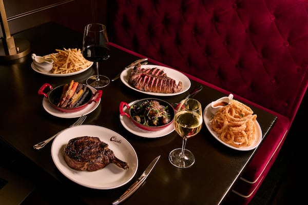 A table full of food at an upscale steakhouse with a tufted velvet banquette, two steaks, onion rings, fries and two glasses of wine