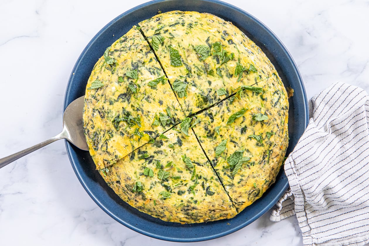 A spinach frittata is on a serving plate. It is cut into 4 pieces, with a silver pie server tucked under one of the pieces. The plate is on a marble tabletop, and a dishcloth is arranged next to it.