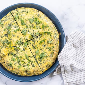 A spinach frittata is on a serving plate. It is cut into 4 pieces, with a silver pie server tucked under one of the pieces. The plate is on a marble tabletop, and a dishcloth is arranged next to it.