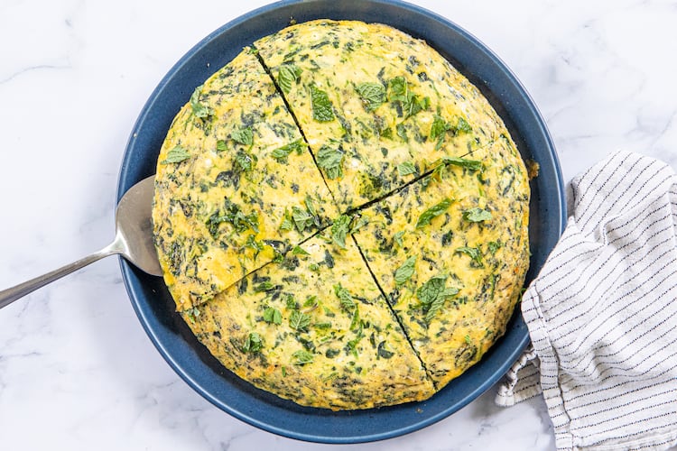 A spinach frittata is on a serving plate. It is cut into 4 pieces, with a silver pie server tucked under one of the pieces. The plate is on a marble tabletop, and a dishcloth is arranged next to it.