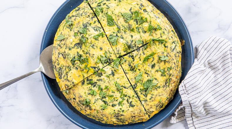 A spinach frittata is on a serving plate. It is cut into 4 pieces, with a silver pie server tucked under one of the pieces. The plate is on a marble tabletop, and a dishcloth is arranged next to it.