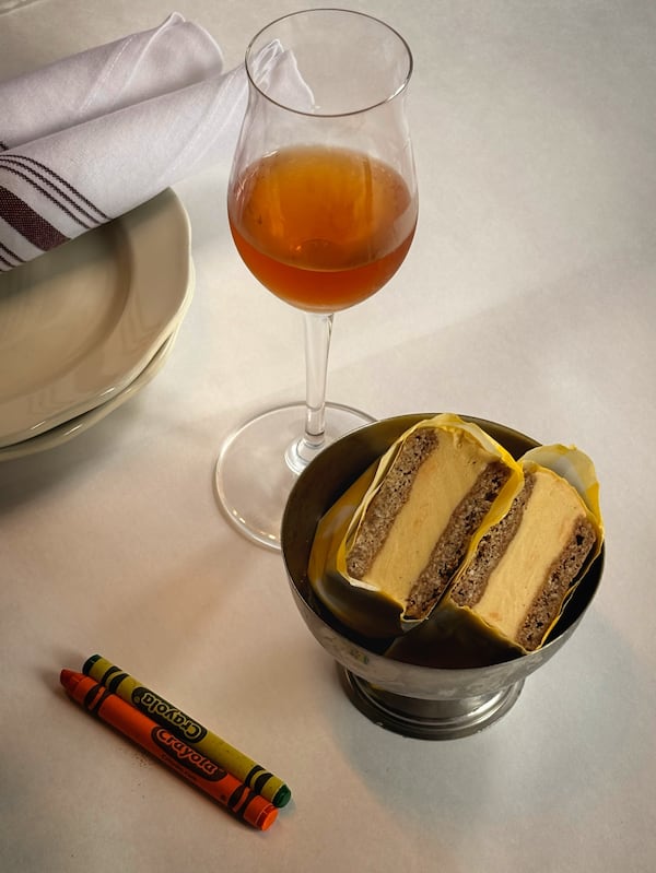A homemade ice cream sandwich with paw paw ice cream is wrapped in paper and served in halves in a small, silver bowl. A glass of orange wine stands next to it.