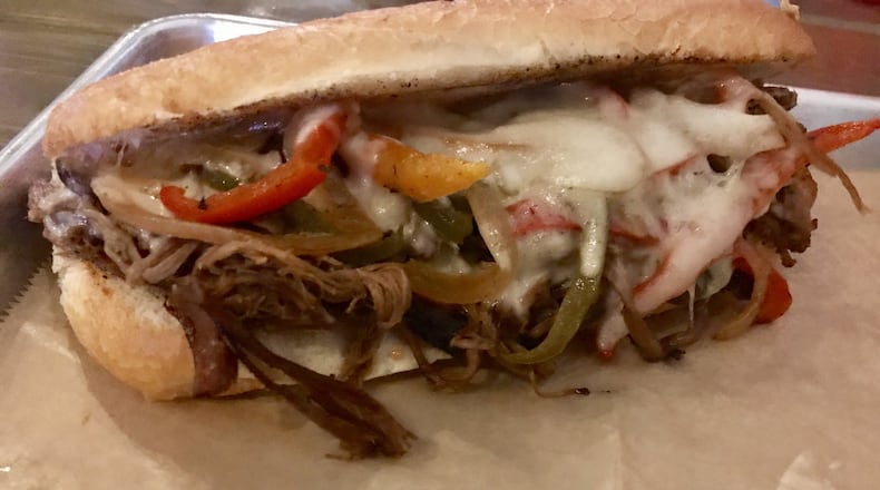 The Philly French Dip at Mouth of the South features smoked brisket, a colorful trio of grilled pepper slices and onions, gooey melted Swiss and provolone cheeses and Sriracha mayo on a toasted hoagie. The sandwich is served with au jus and comes with a side of chips and a pickle. LIGAYA FIGUERAS / LFIGUERAS@AJC.COM