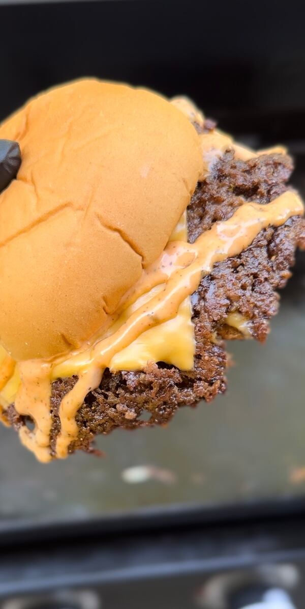 A gloved hand presents a large double smash burger on a plain bun. The melted American cheese is dripping off the side.