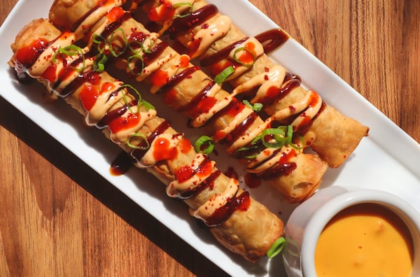 A serving tray displays three eggroll taquitos and dipping sauce. The rolls are drizzled with sauces and garnished with chopped green onions.