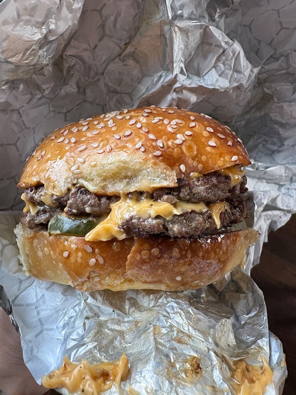 Two burger patties with a slice of melted cheese inbetween are on a thick sesame seed hamburger bun. The burger is shown in a foil wrapper, with cheese stuck to the wrapper in three spots.
