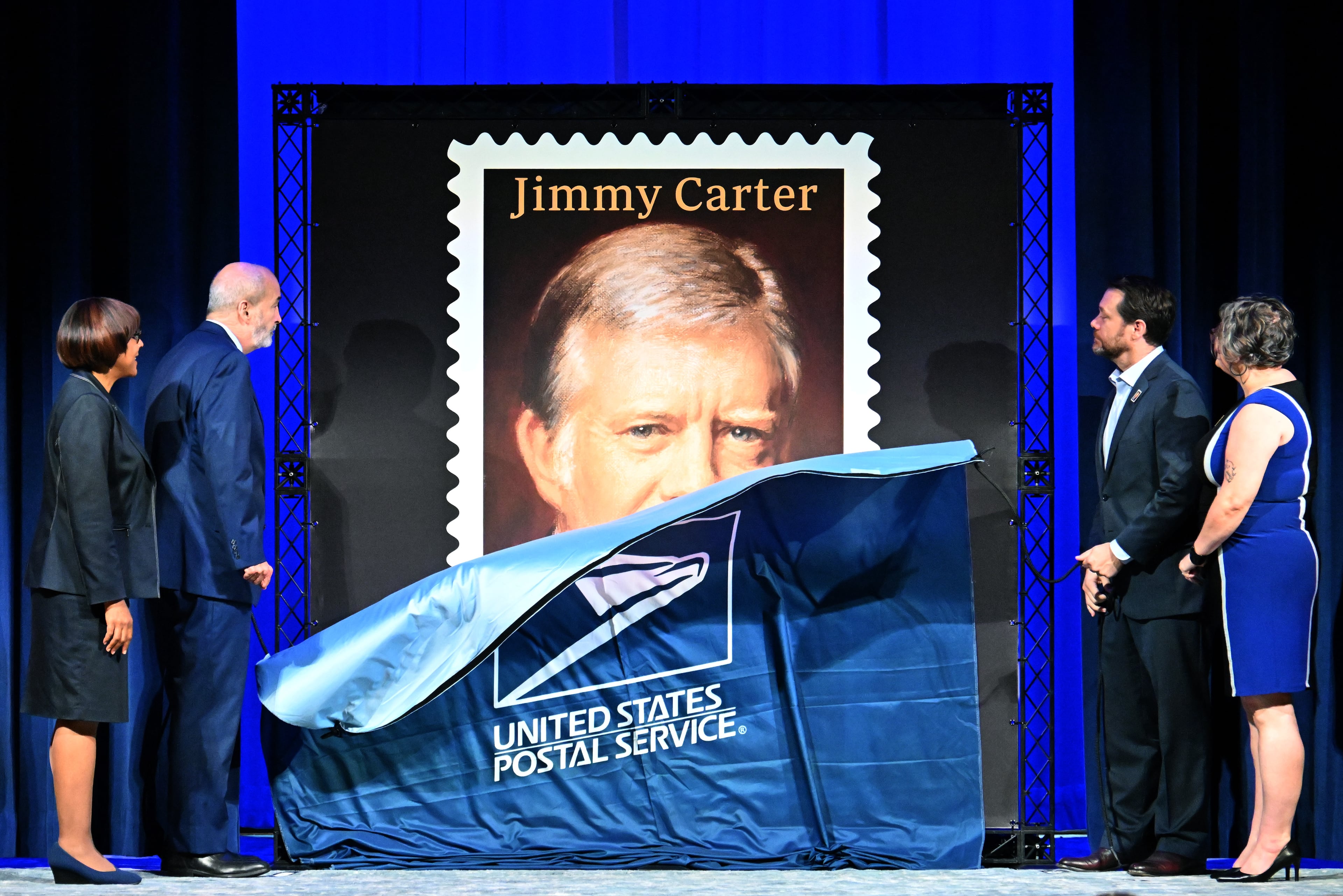 Crystal Edmonson, broadcast dditor of Atlanta Business Chronicle (from left); Thomas Marshall, general counsel of USPS; Jason Carter, grandson of former President Jimmy Carter; and Beth Davis, chief operating officer of the Carter Center, unveil the Jimmy Carter "forever" stamp in Atlanta on Wednesday. (Hyosub Shin/AJC)