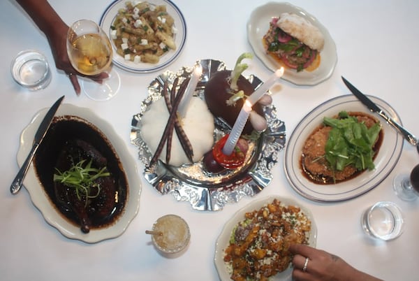 An overhead view of several plates of food on a white tablecloth. Starting bottom left and moving clockwise, there's a plate of chicken covered in a dark sauce, a wine glass, a plate of oil-cured eggplant, a sandwich, a plate of schnitzel and a salad.
