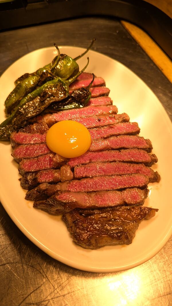 A plate filled with slices of rare boneless ribeye steak, topped with an egg yolk. The steak is garnished with four full-sized grilled peppers.