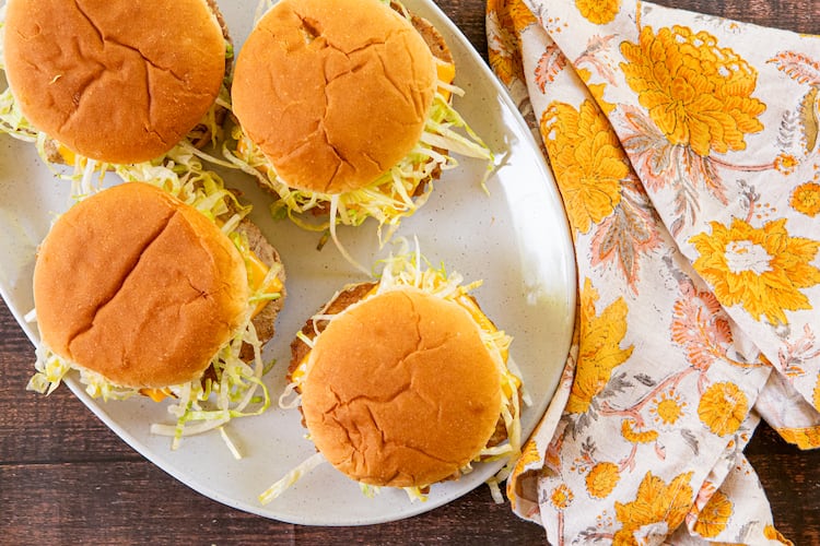 A platter of four turkey smash burgers, topped with shredded lettuce and American cheese, is shown with a floral cloth napkin beside it.