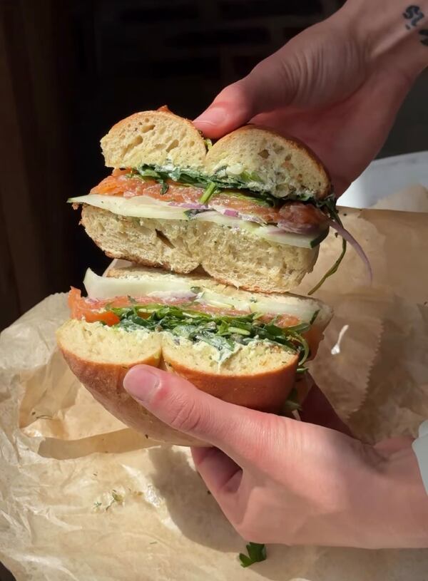 Two hands hold a gravlax bagel sandwich cut in two, opening the sandwich so the cut sides are visible to the camers