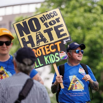 A small group of protestors rally before a school board meeting in Decatur on Thursday. SJ, his activist name, wears stickers and a T-shirt showing him as a “safe zone.” Protesters are upset with what they believe is a lack of transparency around building an early learning center and the district's connection to the superintendent's controversial podcaster husband. (Estela Muñoz/AJC)