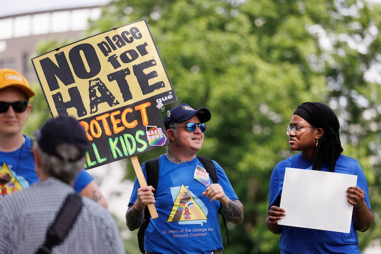 A small group of protestors rally before a school board meeting in Decatur on Thursday. SJ, his activist name, wears stickers and a T-shirt showing him as a “safe zone.” Protesters are upset with what they believe is a lack of transparency around building an early learning center and the district's connection to the superintendent's controversial podcaster husband. (Estela Muñoz/AJC)