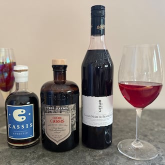 Three bottles of cassis liqueur with a wine glass holding a Kir cocktail to the right