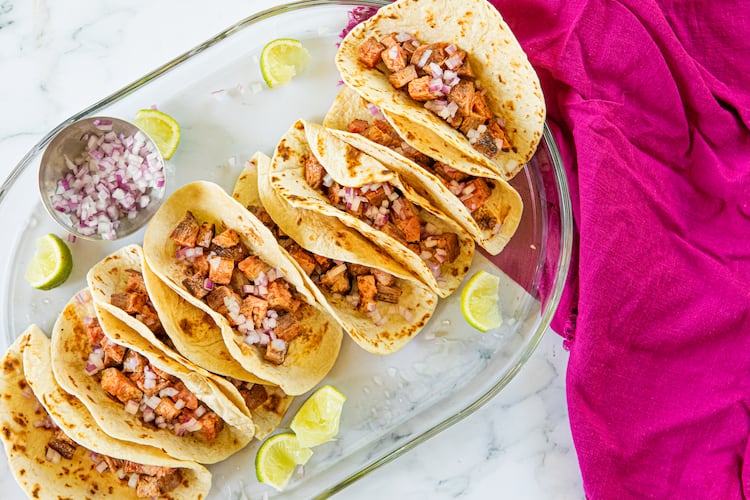 A tray holds 8 carne asada tacos on grilled flour tortillas, topped with diced onions and garnished with lime wedges. A bright towel adds visual interest to the photo.
