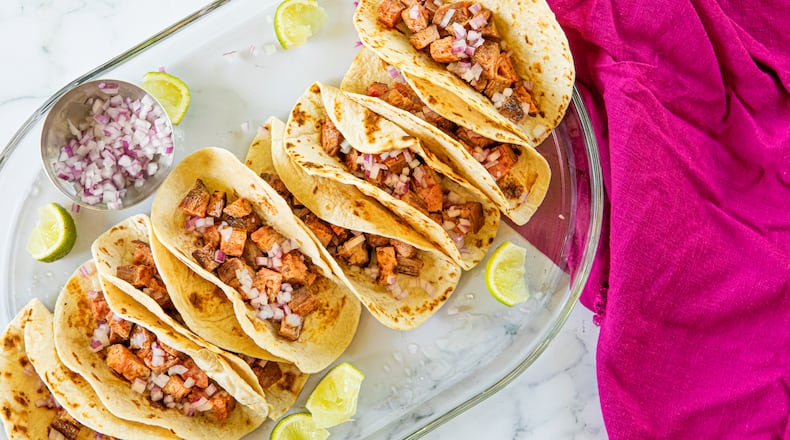 A tray holds 8 carne asada tacos on grilled flour tortillas, topped with diced onions and garnished with lime wedges. A bright towel adds visual interest to the photo.
