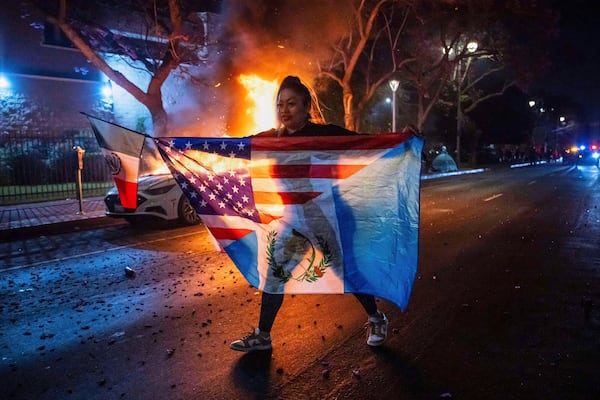 A person displaying multiple flags walks past a burning car during protests over the Trump administration's immigration raids in Los Angeles, Monday, June 9, 2025. (AP Photo/Ethan Swope)
