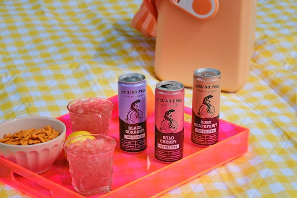 Three cans of Cycling Frog beverages are arranged on a lucite tray, which also holds a bowl of snack mix and two rocks glasses filled with ice and the beverages. The tray rests on a gigham tablecloth, signaling outdoor entertaining.