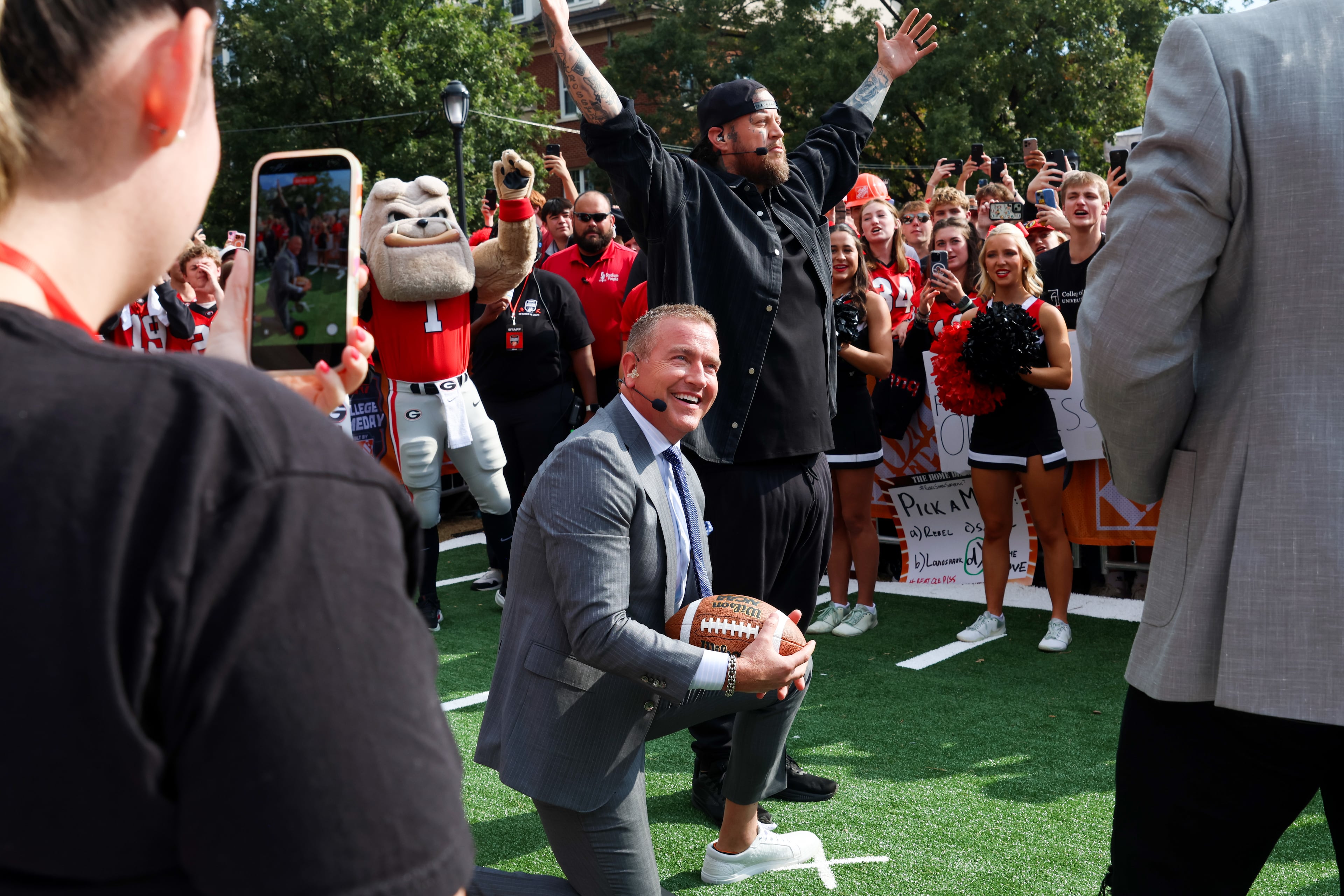 Kirk Herbstreit (kneeling) sets up a ball for country artist Jelly Roll, the week’s guest picker, during Pat McAfee’s Kicking Contest on the ESPN College GameDay broadcast from Myers Quad at UGA on Saturday, Oct. 18, 2025. (C.J. Bartunek for the AJC)