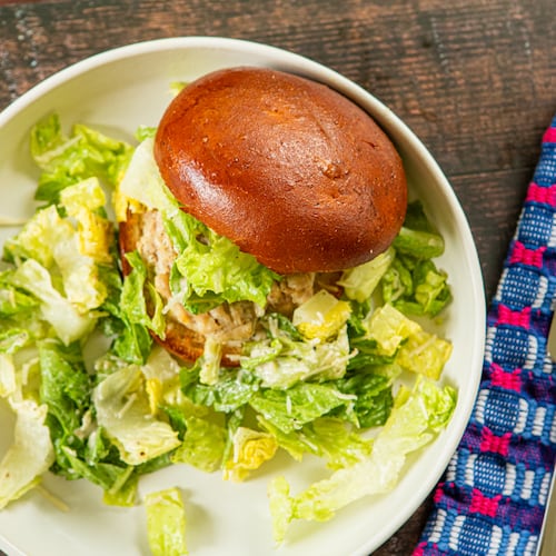 A plate filled with a chicken burger on a smooth bun garnished with Caesar salad, and also with Caesar salad on the side. The plate is on a wooden table top with a fork, knife and napkin next to it.